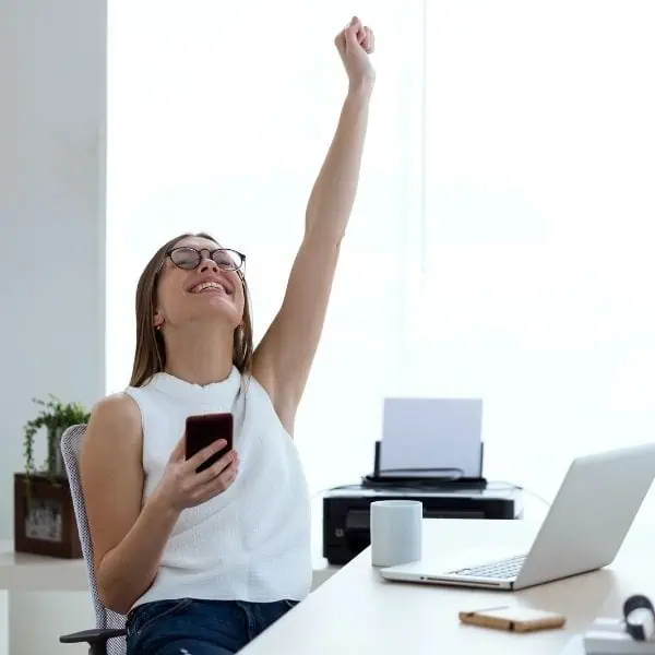 A happy young woman in an office looking upwards with her hand in the air and holding her mobile phone